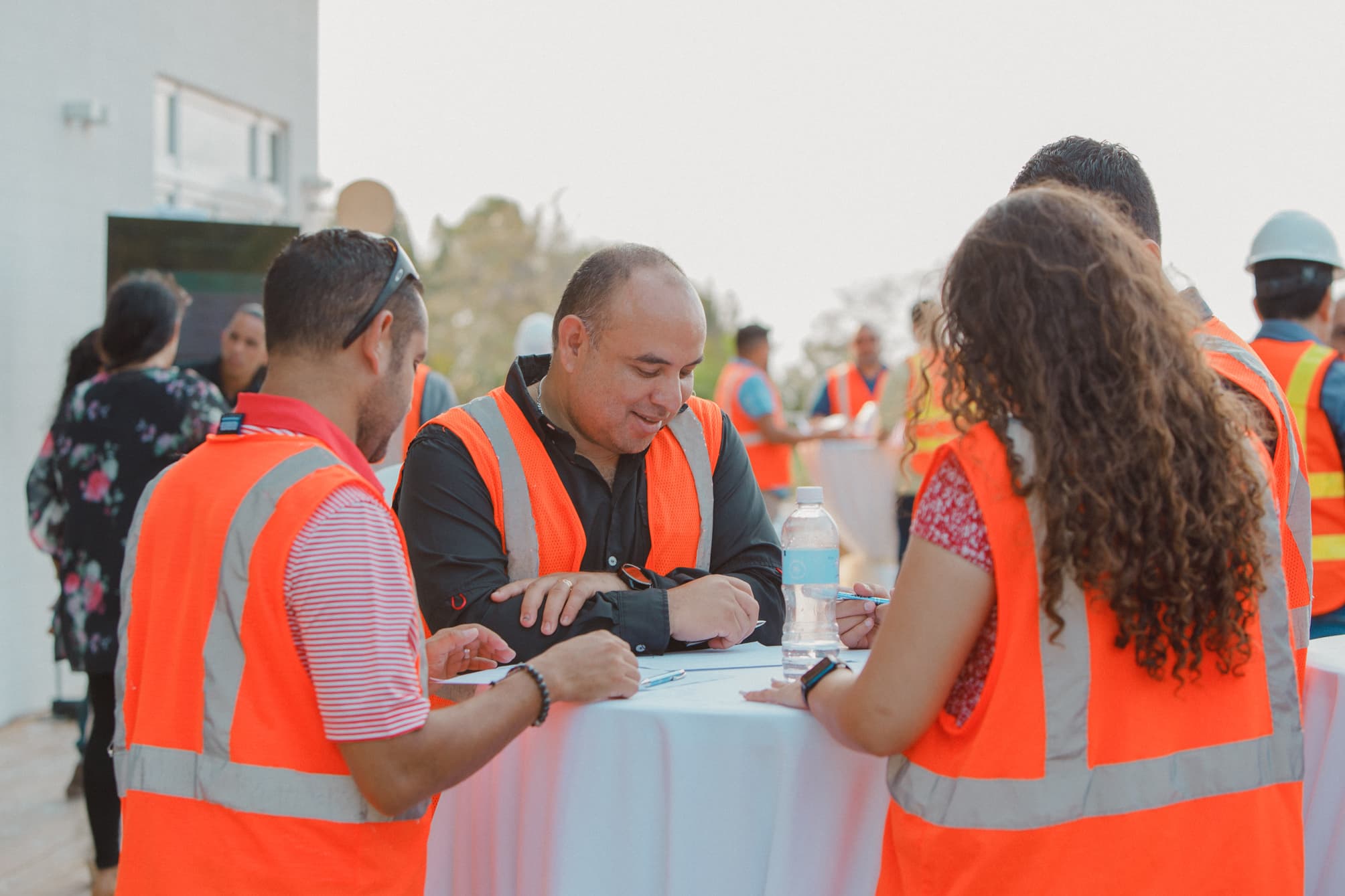 A group of people wearing orange safety vests, gathered around a table and engaged in discussion or planning. The setting appears to be outdoors, with some participants holding documents and others interacting in the background, suggesting a collaborative work environment.