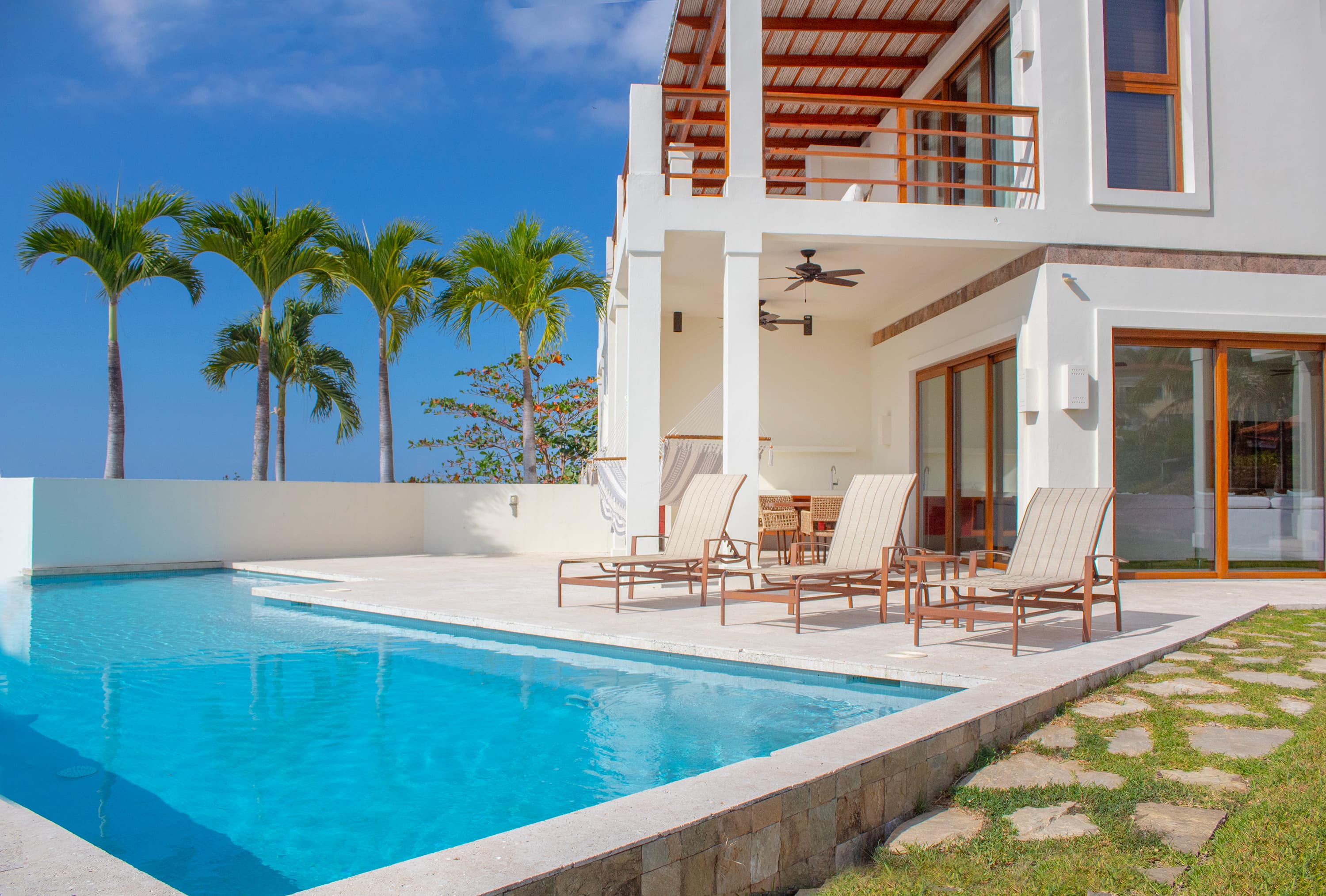 Luxury beachfront villa with private pool, featuring a white modern exterior, spacious patio with lounge chairs, and palm trees against a clear blue sky. Ideal tropical vacation rental property showcasing upscale amenities and indoor-outdoor living.