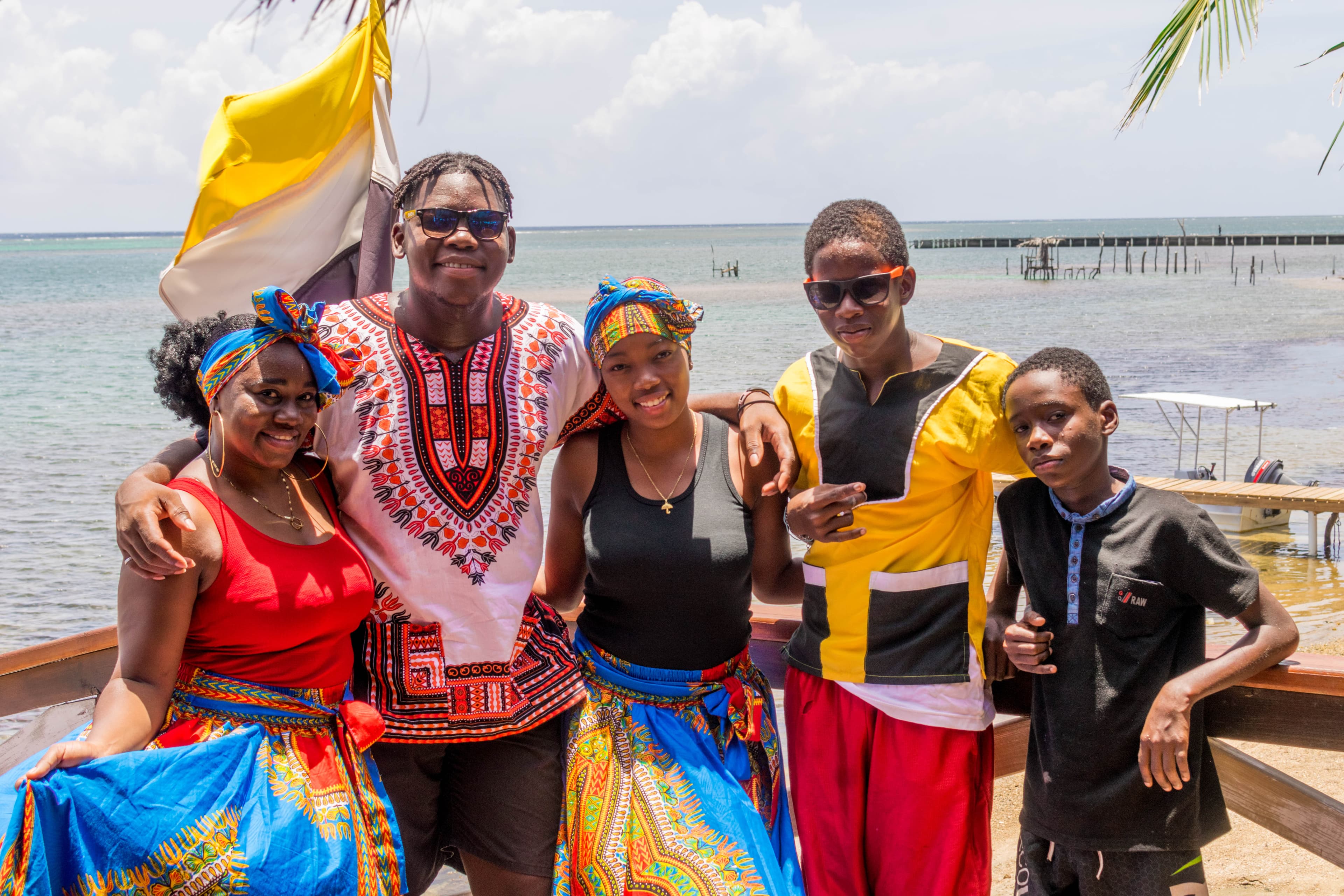 A group of five people standing together on a wooden deck by the water, wearing colorful traditional clothing and smiling. The background features a calm sea with a dock and a partly cloudy sky, suggesting a festive or cultural gathering.